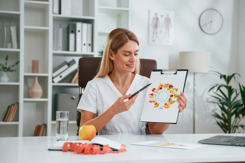 With picture of food and vitamins. Young female doctor in white coat is indoors